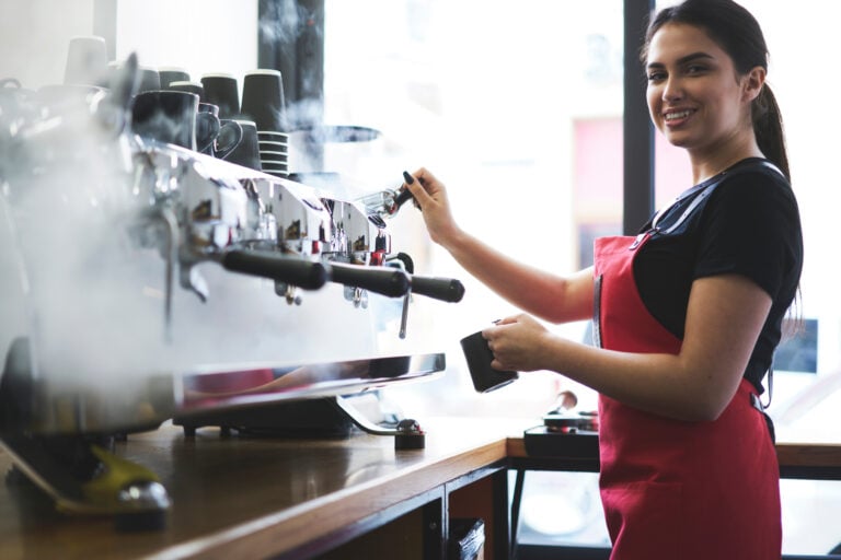 Smiling busty female barista using professional coffee machine enjoying job in cafe improving skills of making cappuccino